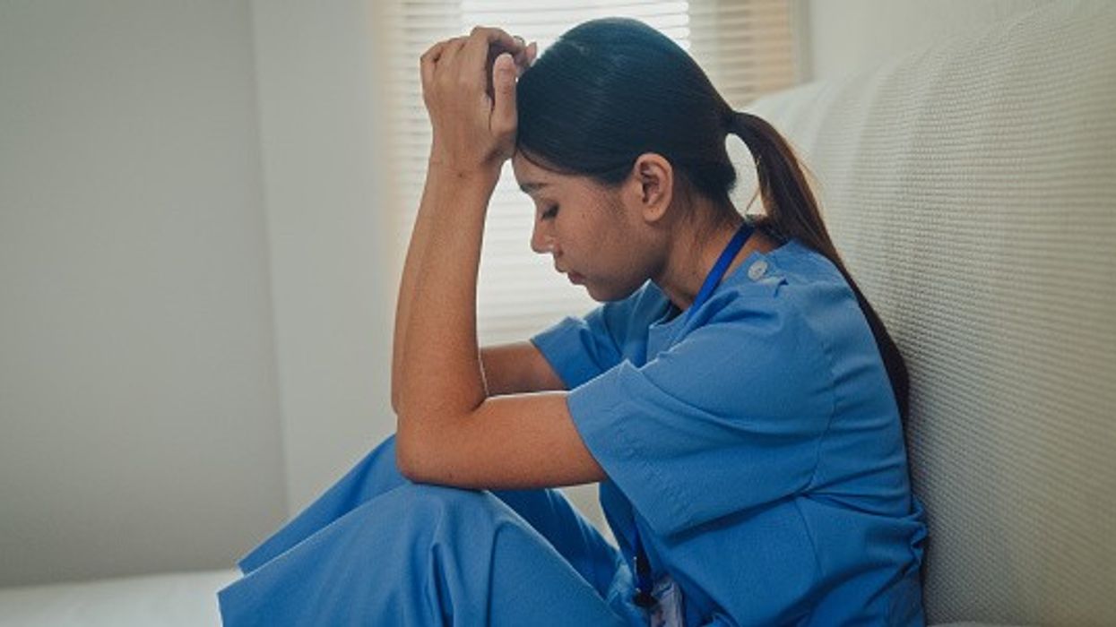 Stressed NHS nurse in uniform sits with head in hands amid job loss fears and budget cuts.